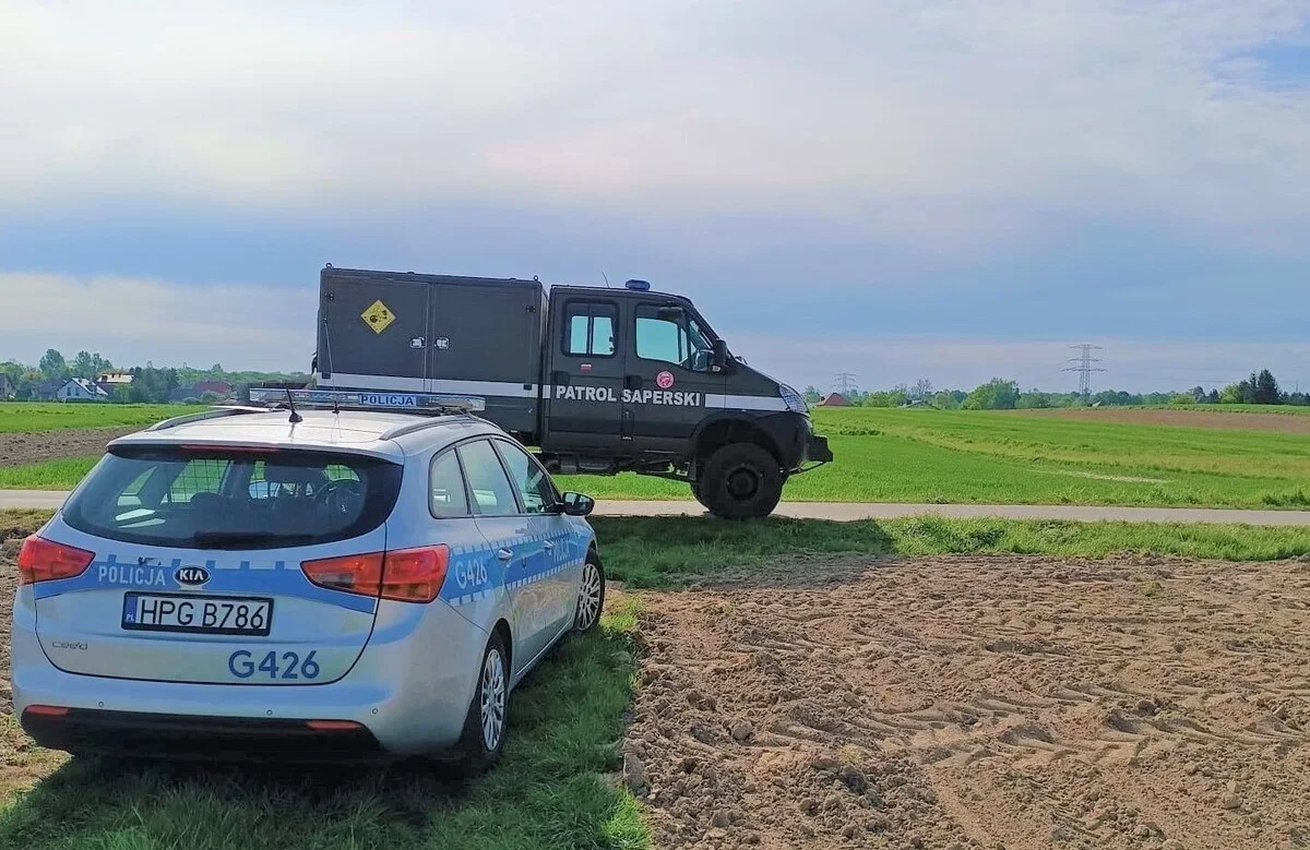 Policyjny radiowóz i pojazd patrolu saperskiego stoją przy polnej drodze w otoczeniu pól.