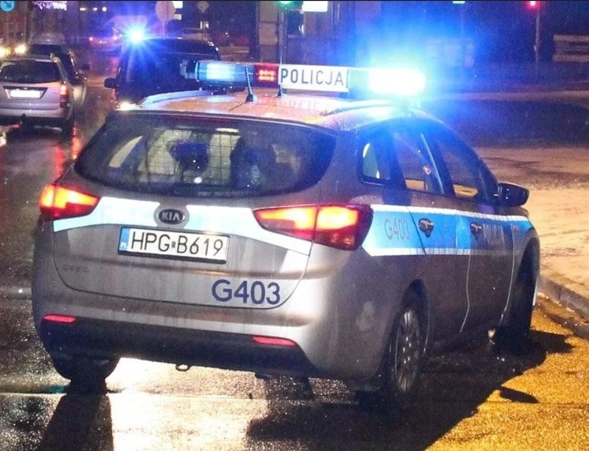 Policyjny radiowóz Kia z oznaczeniem G403 i napisem POLICJA stoi nocą na mokrej ulicy miejskiej z włączonymi niebieskimi światłami błyskowymi.