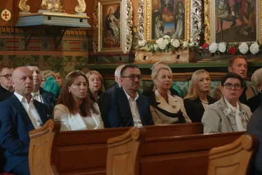 Grupa osób siedzi na drewnianych ławkach w bogato zdobionym wnętrzu kościoła, w tle widoczne ołtarzowe obrazy i kwiaty.