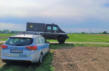 Policyjny radiowóz i pojazd patrolu saperskiego stoją przy polnej drodze w otoczeniu pól.