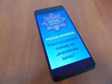 Smartfon leży na drewnianym blacie i wyświetla ostrzeżenie policji z Oświęcimia przed oszustwami metodą na pracownika banku.