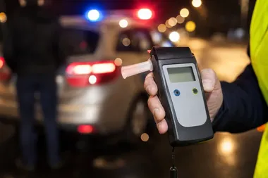 Funkcjonariusz w odblaskowej kamizelce trzyma alkomat podczas nocnej kontroli drogowej z radiowozem w tle.