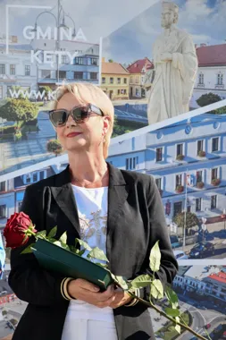 Kobieta w czarnej marynarce i białej bluzce trzyma czerwoną różę i zieloną teczkę, stojąc przed banerem promującym Gminę Kęty.